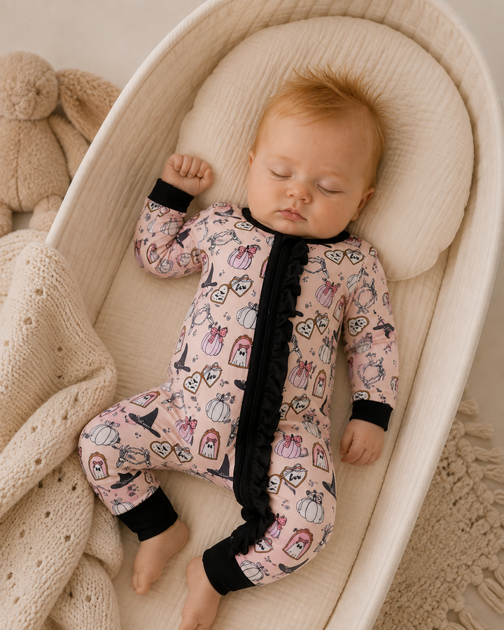 A sleeping baby wearing a pink full-body pajama with a black ruffled zipper and black cuffs, lying on a cream bassinet with a knitted blanket and a teddy bear nearby.