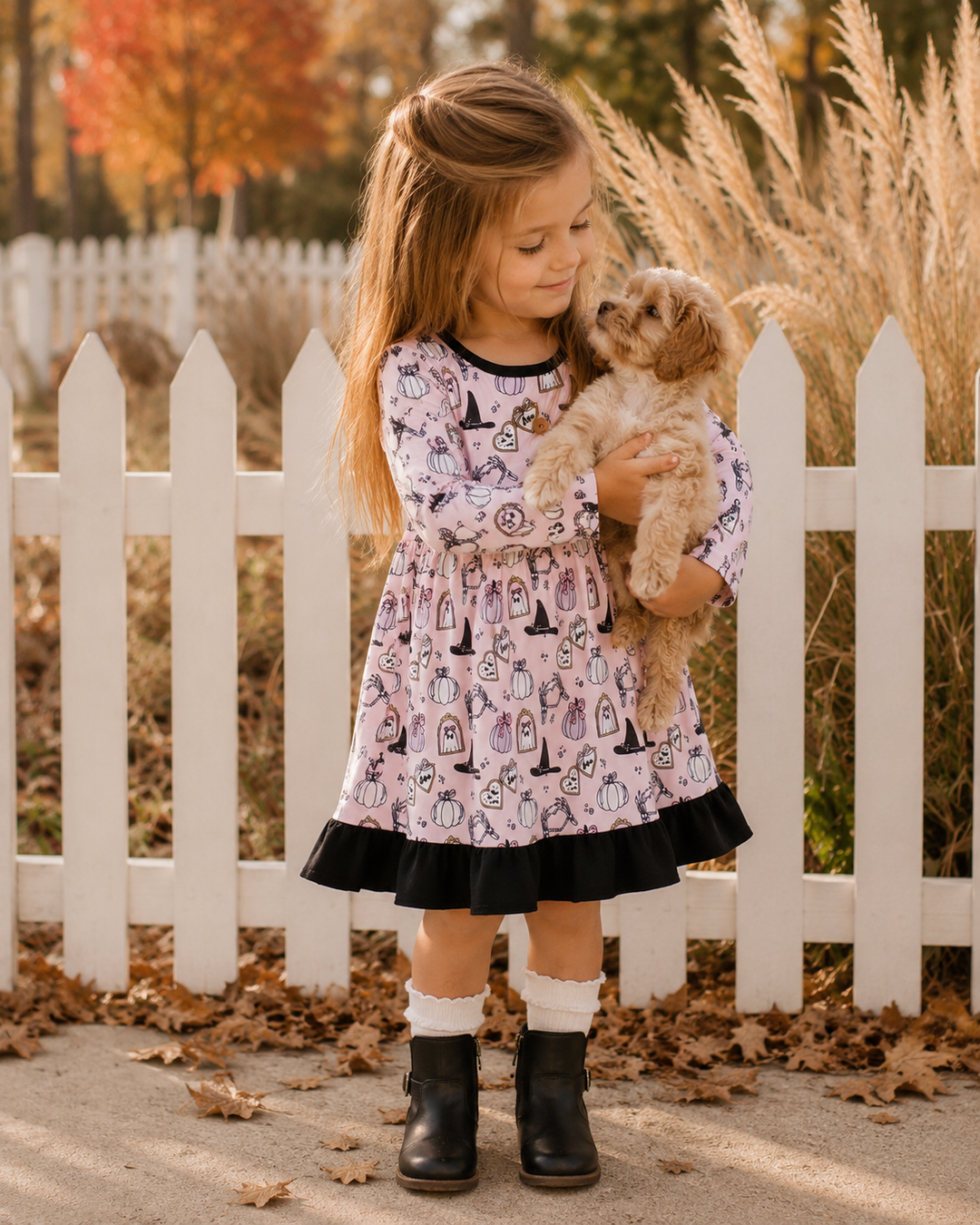 A young girl stands outdoors in front of a white picket fence holding a small fluffy dog. She is wearing a light pink long-sleeve dress with a black and white witch-themed print, a black ruffled hem, white socks, and black ankle boots.