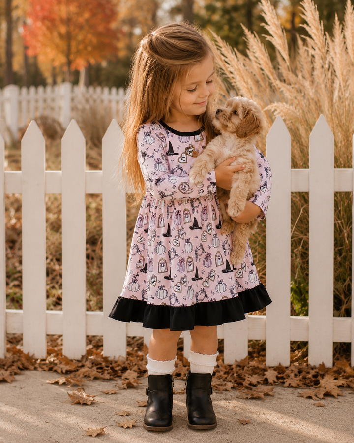 A young girl stands outdoors in front of a white picket fence holding a small fluffy dog. She is wearing a light pink long-sleeve dress with a black and white witch-themed print, a black ruffled hem, white socks, and black ankle boots.