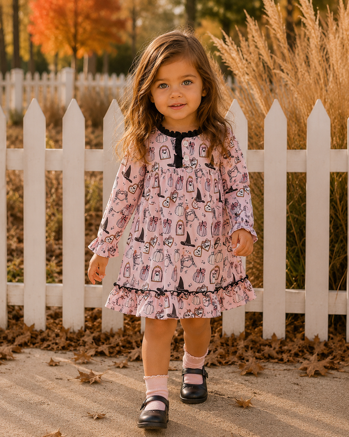 A young girl stands outdoors in front of a white picket fence wearing a pink long-sleeve dress with a black ruffled collar and hem, an allover witch-themed print, pink socks, and black Mary Jane shoes.