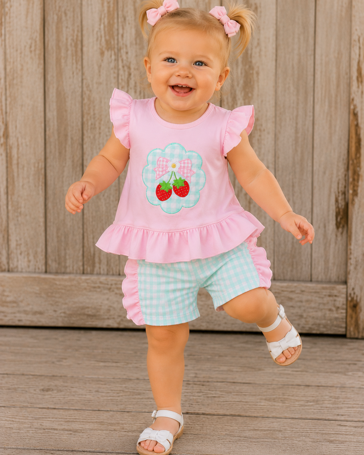Child wearing a pink ruffled top with cherry design and light blue shorts on a wooden floor.