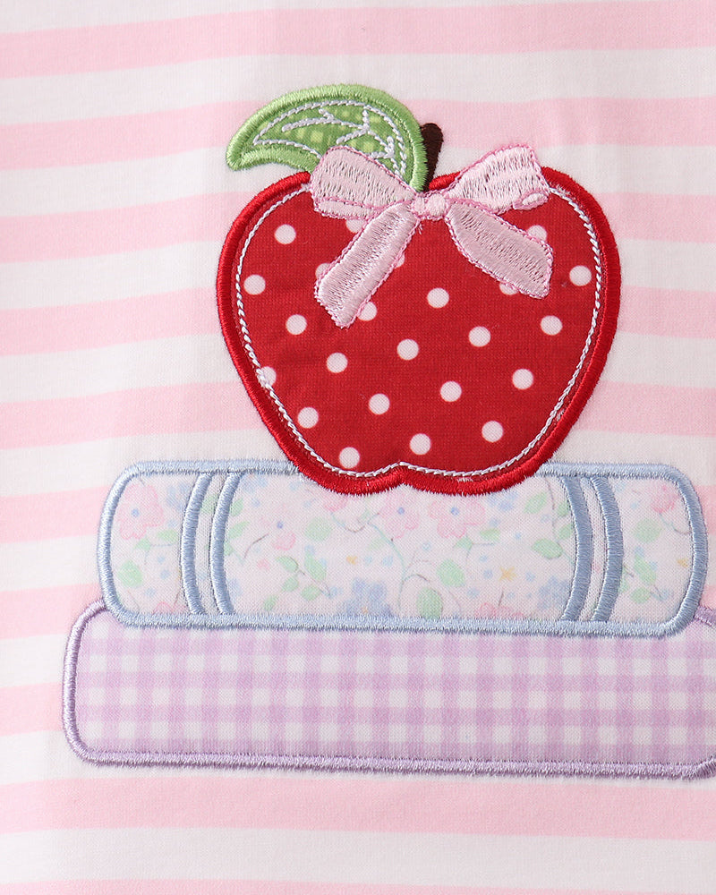 A close-up of a pink and white striped fabric featuring an applique design of a red apple with white polka dots, a green leaf, a pink bow, and two pastel patterned book shapes underneath.