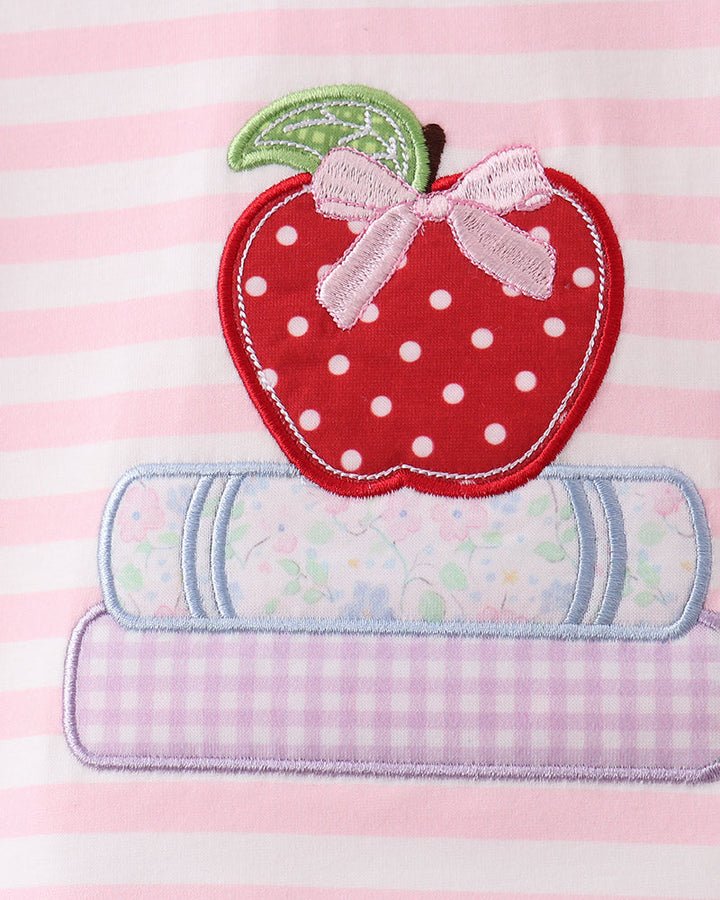 A close-up of a pink and white striped fabric featuring an applique design of a red apple with white polka dots, a green leaf, a pink bow, and two pastel patterned book shapes underneath.