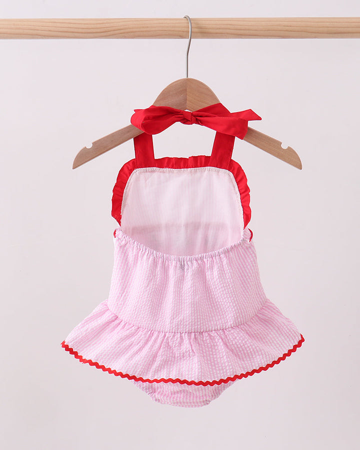 Pink baby dress with red trim and bow on a hanger against a white background