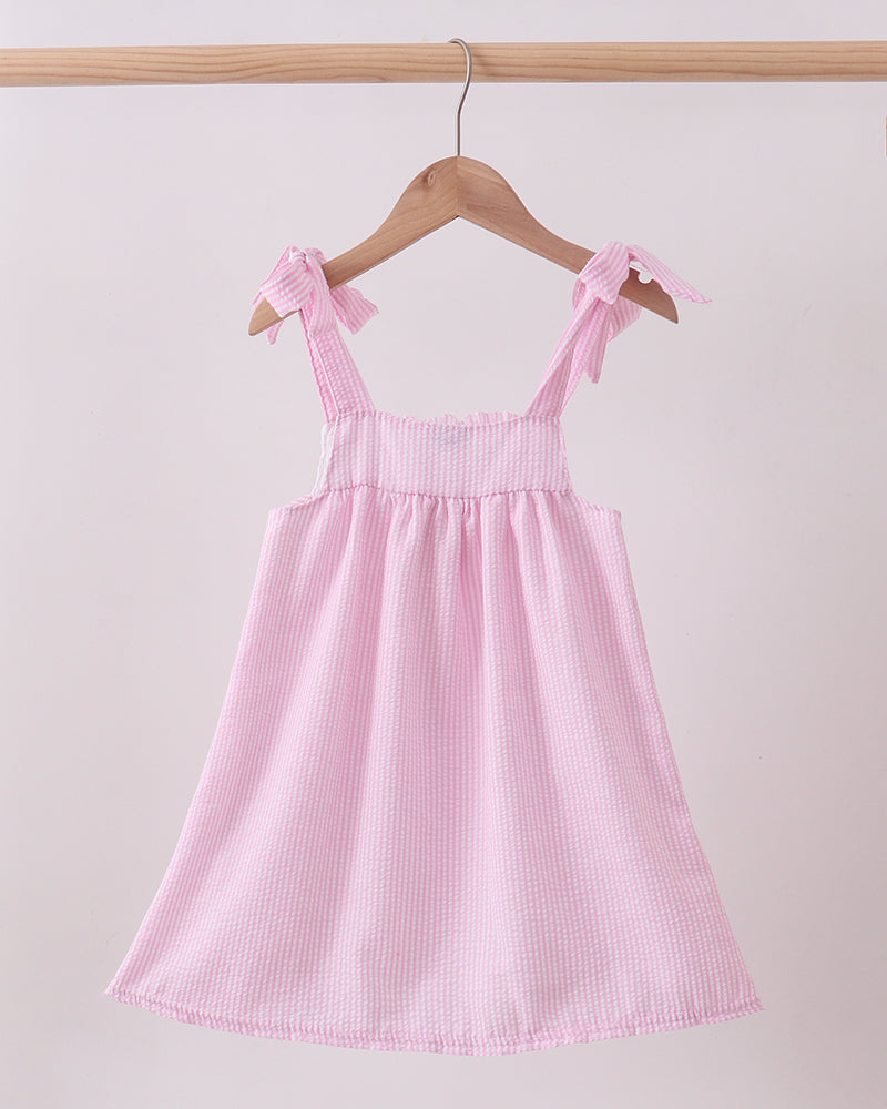 Pink dress with bow straps hanging on a wooden hanger against a light pink background