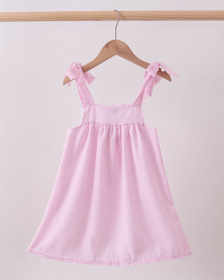Pink dress with bow straps hanging on a wooden hanger against a light pink background