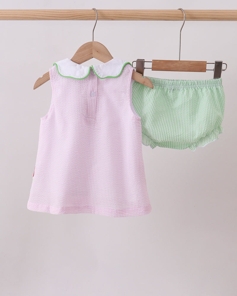 Pink dress with green trim and matching green bloomers hanging on wooden hangers against a light gray background.
