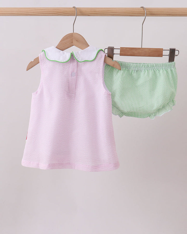 Pink dress with green trim and matching green bloomers hanging on wooden hangers against a light gray background.