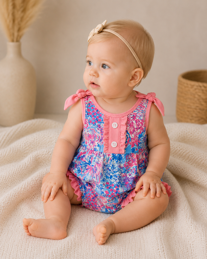 Baby wearing a colorful floral romper with pink accents, sitting on a beige blanket.