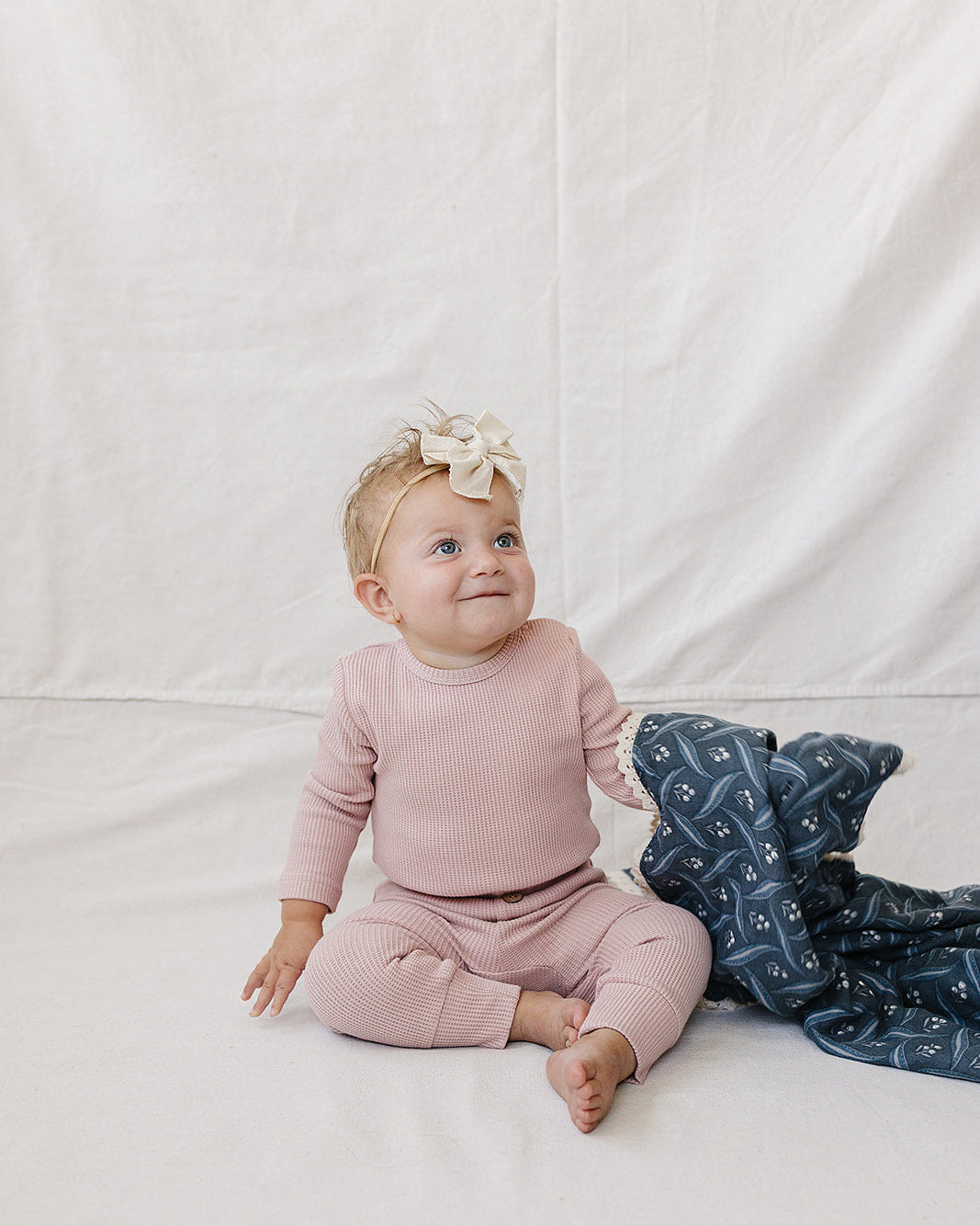 A baby sitting on a white fabric backdrop wearing a dusty pink long-sleeve top and matching pants, with a cream bow in their hair and a navy blanket with a white pattern beside them.