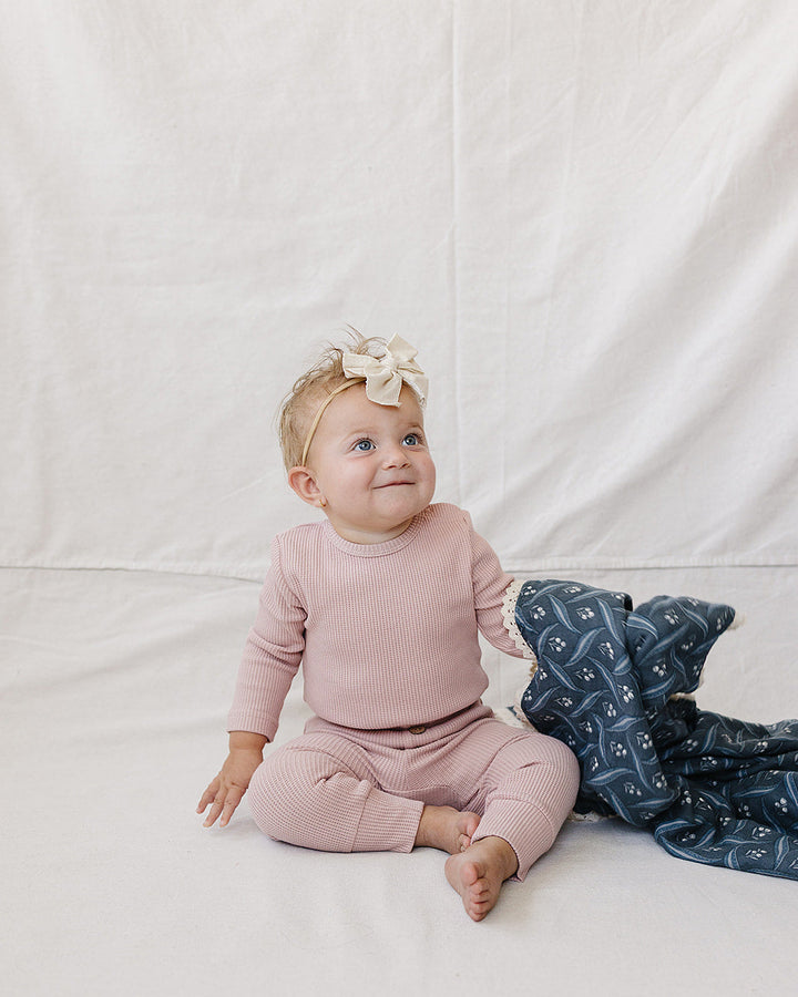 A baby sitting on a white fabric backdrop wearing a dusty pink long-sleeve top and matching pants, with a cream bow in their hair and a navy blanket with a white pattern beside them.
