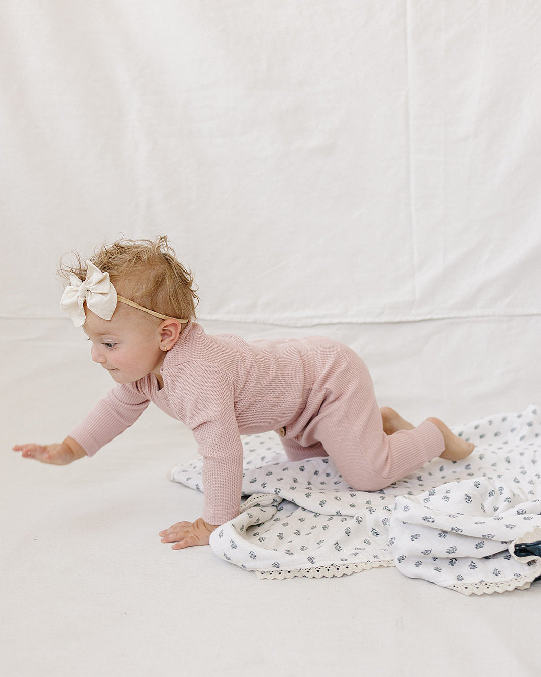 A baby crawling on a white patterned blanket, wearing a dusty pink long-sleeve romper and a white bow headband, set against a white fabric backdrop.