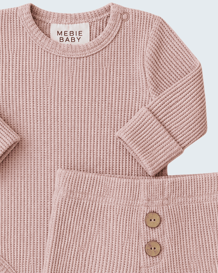 A close-up of a baby knit two-piece outfit in dusty pink, featuring a long-sleeve crewneck top and matching pants with two wooden buttons, from the brand MEBIE BABY.
