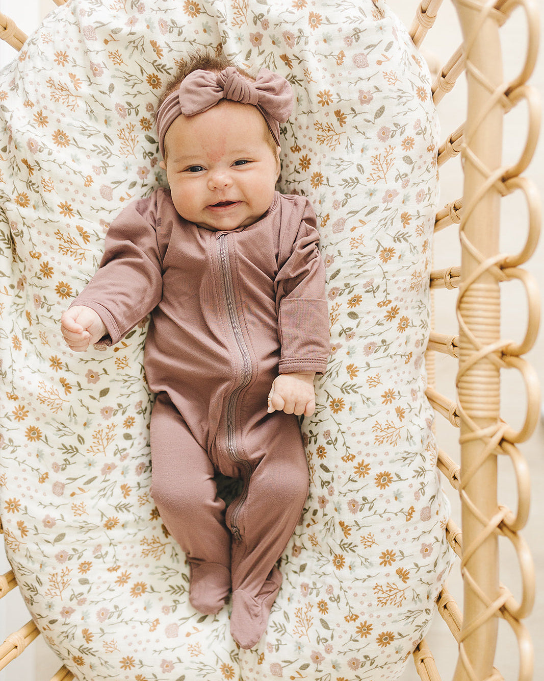 Baby in a pink outfit lying on a floral-patterned cushion inside a wooden crib.