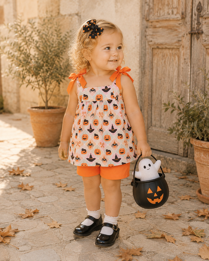A toddler girl stands outdoors in a rustic courtyard wearing a sleeveless Halloween dress with a pumpkin and witch print, orange shorts, white socks, and black Mary Jane shoes. She holds a black jack-o&