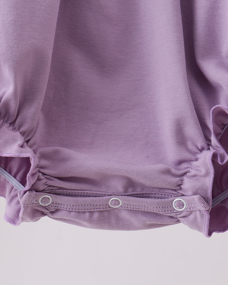 Close-up of a baby romper in dusty lavender purple, showing the gathered ruffled hem with three silver metal snap buttons on a light gray background.