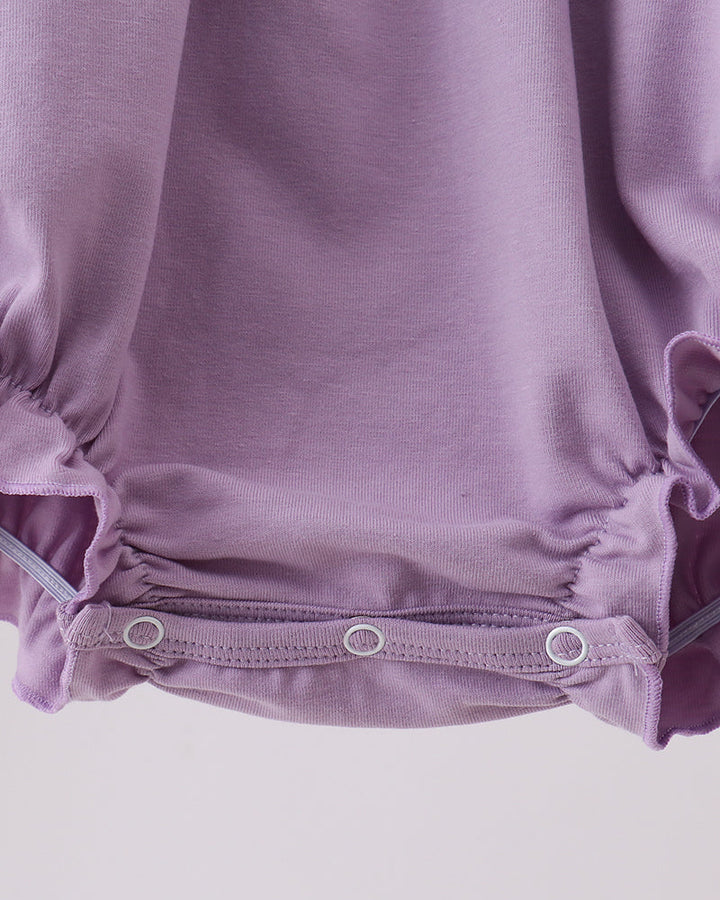 Close-up of a baby romper in dusty lavender purple, showing the gathered ruffled hem with three silver metal snap buttons on a light gray background.