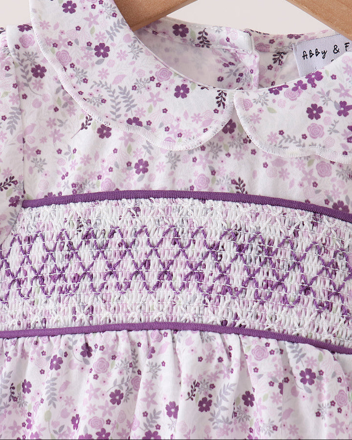 A close-up of a baby romper on a wooden hanger, featuring a white base with a purple and lavender floral pattern, a smocked chest panel, and a Peter Pan collar.