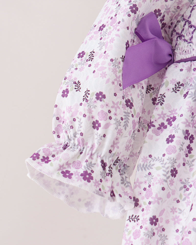 A close-up of a white garment with a purple and pink floral print, featuring a large purple bow at the shoulder and ruffled sleeves, set against a plain light beige background.