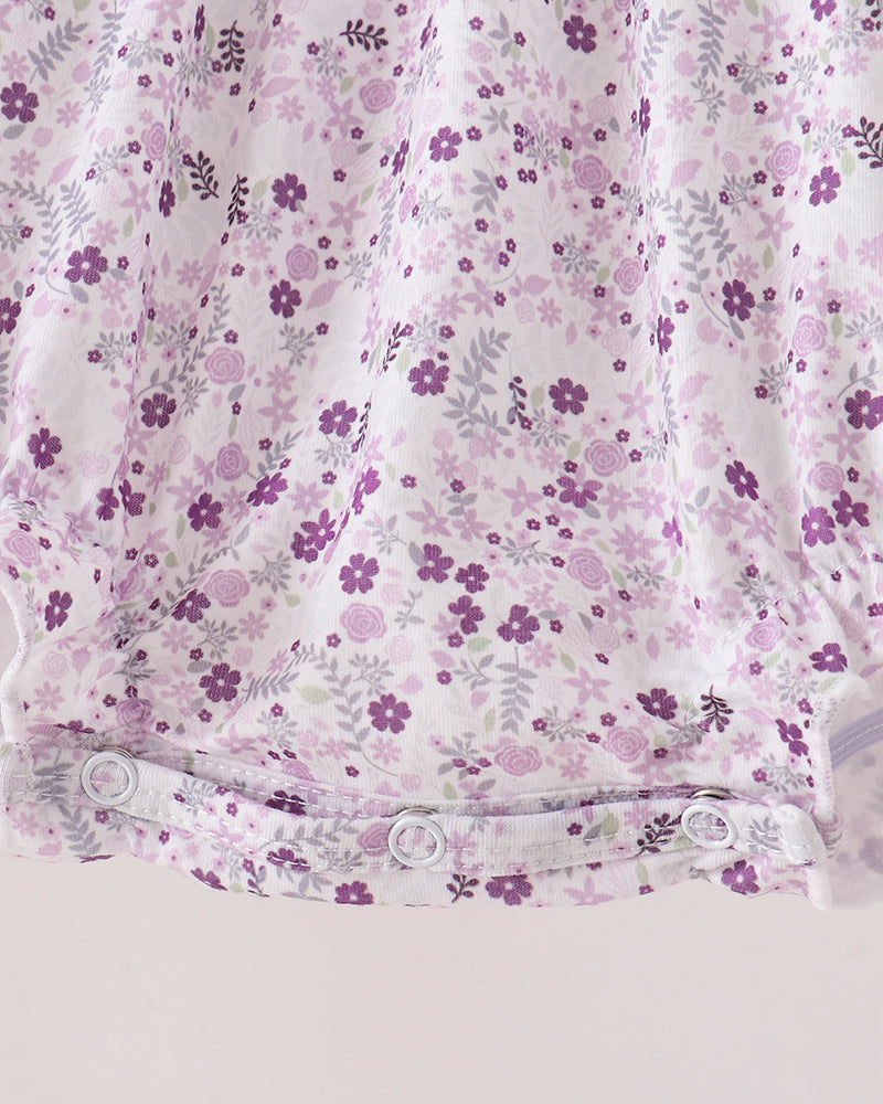 Close-up of a baby garment or fabric panel with a white base and an all-over purple floral print, featuring a snap-button waistband.