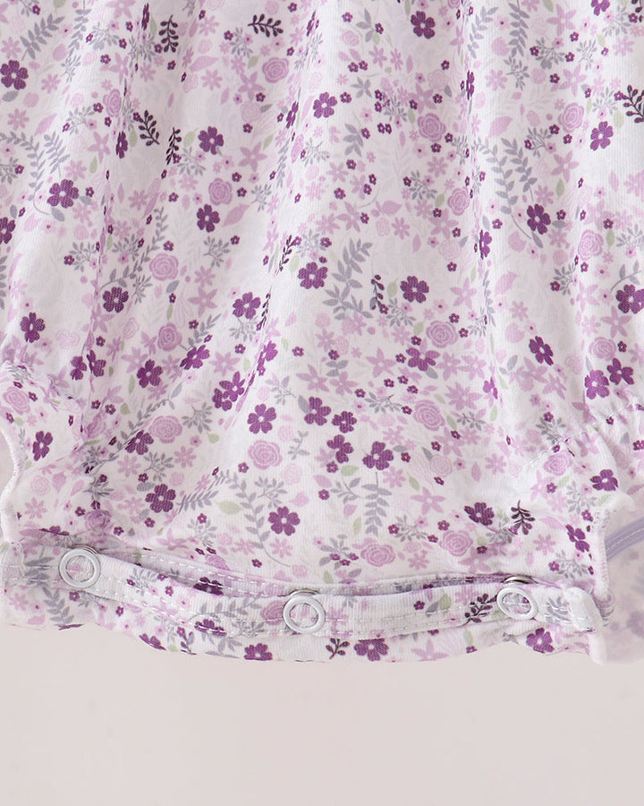 Close-up of a baby garment or fabric panel with a white base and an all-over purple floral print, featuring a snap-button waistband.