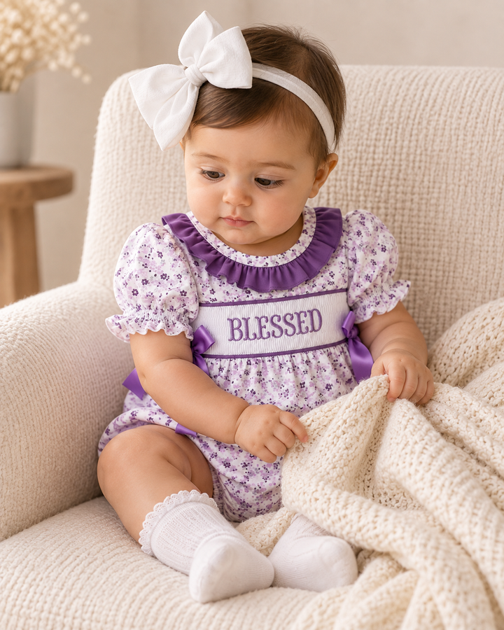 Baby girl in a floral romper and large white bow. She is sitting in a comfy chair.