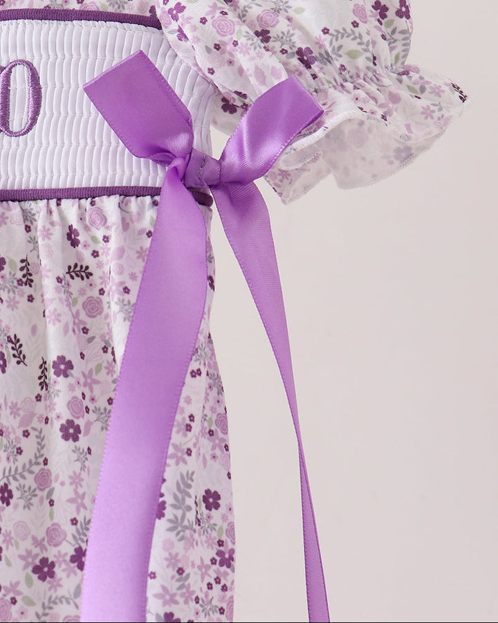 Close-up of a white baby dress with a purple floral print, featuring a white embroidered chest panel with a purple letter &