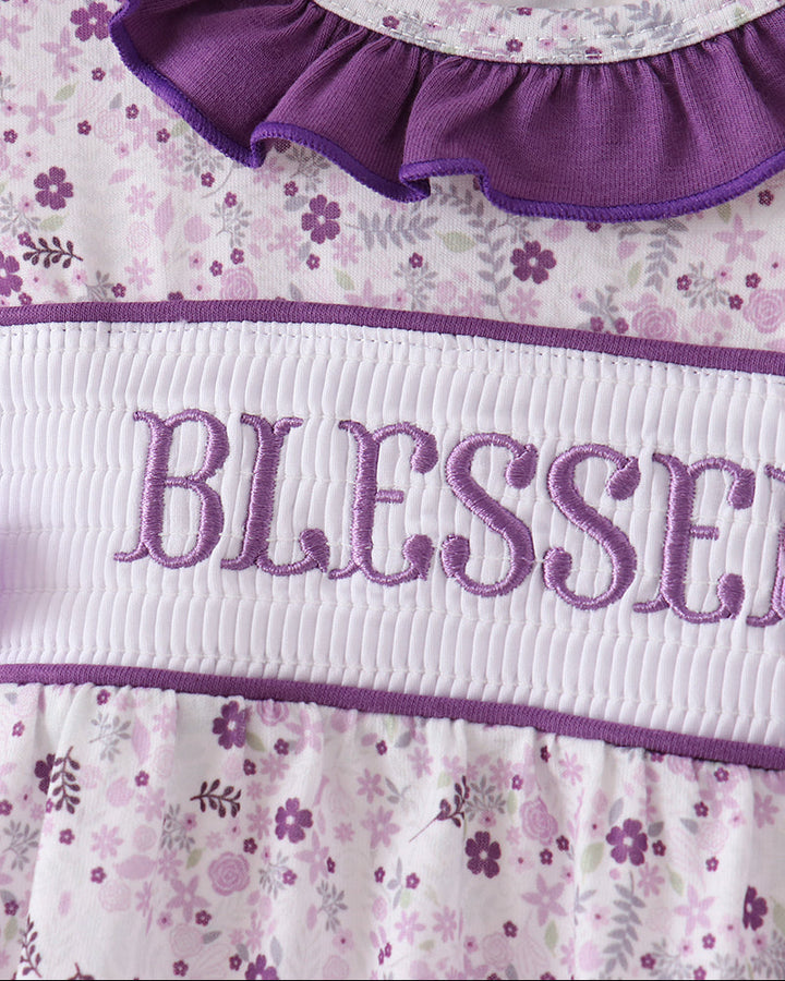 Close-up of a baby dress with a purple ruffled collar, a white smocked band embroidered with &