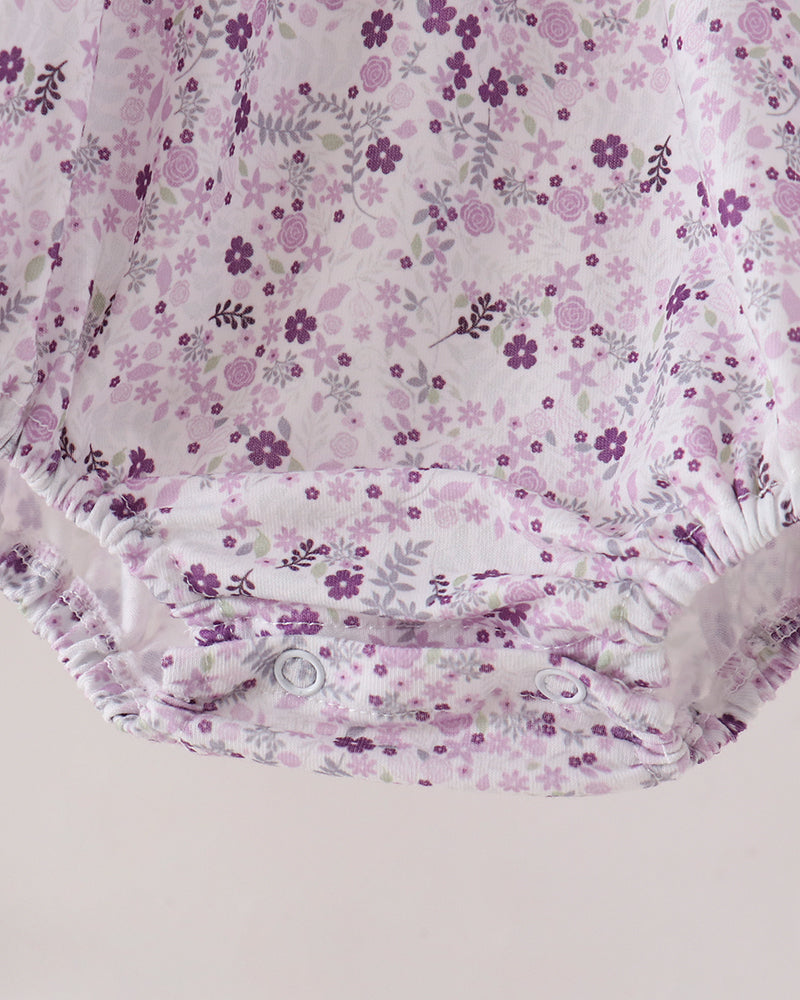 A close-up of a baby romper or bodysuit featuring a delicate purple and lavender floral pattern on a white background, with elasticized snap closures at the bottom.