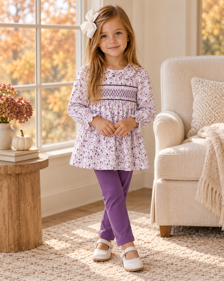 Young girl in a purple floral outfit is standing in front of large window.