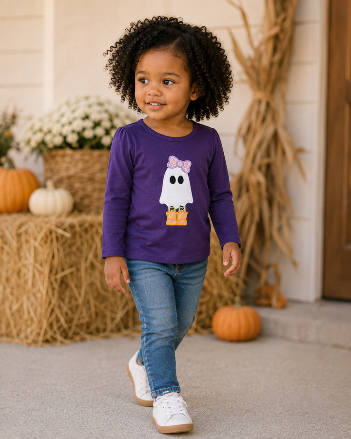 A toddler girl with curly black hair wearing a purple long-sleeve shirt with a white ghost graphic, blue jeans, and white sneakers, standing on a porch decorated with pumpkins and hay bales.