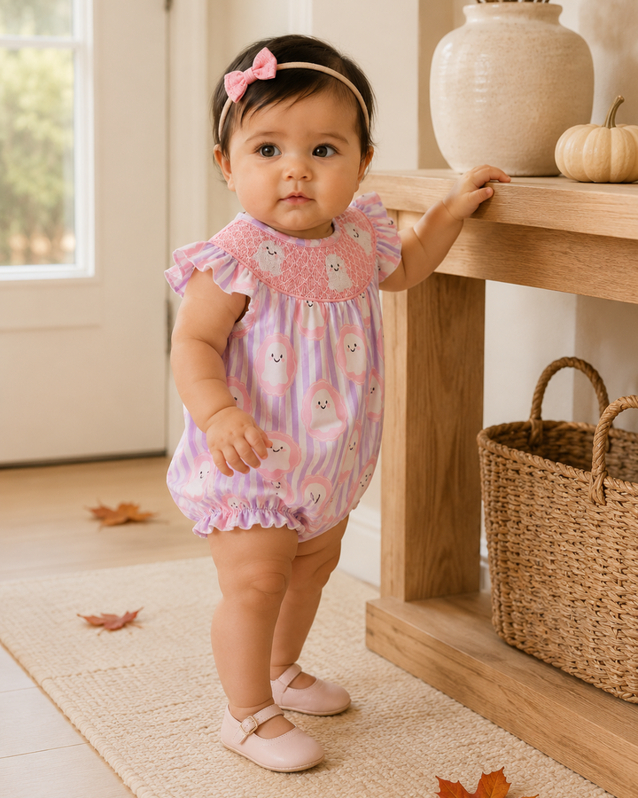 A toddler girl wearing a pink and lavender striped romper with embroidered cartoon faces and a pink bow headband, standing in a warm, autumn-themed room next to a wooden console table.