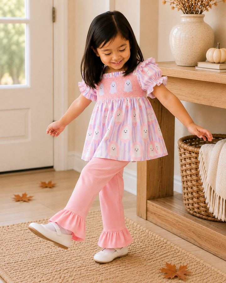 A young girl wearing a pink and white owl-patterned smocked dress with ruffled sleeves and matching pink ruffled pants, standing on a woven rug in a warm, decorated room.