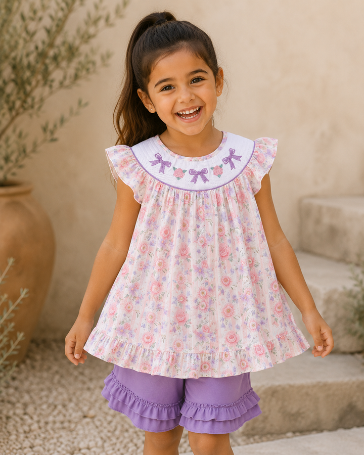 Young girl wearing a floral top and purple shorts with ruffles, standing outdoors.