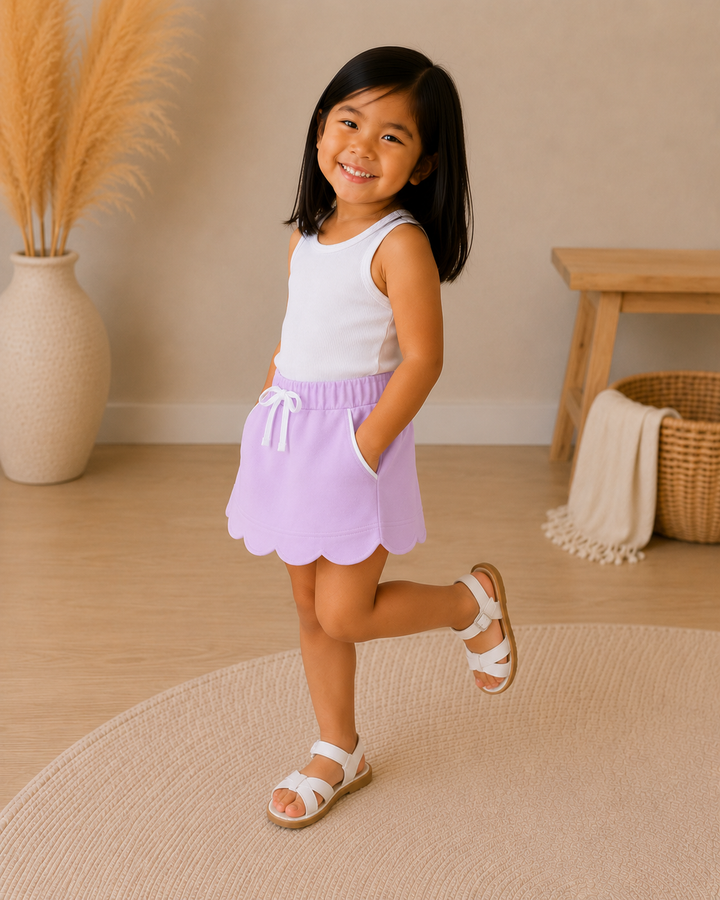 A young girl smiling and posing in a white sleeveless top and a lavender purple scalloped skort with white sandals, set in a warm, neutral-toned indoor room.