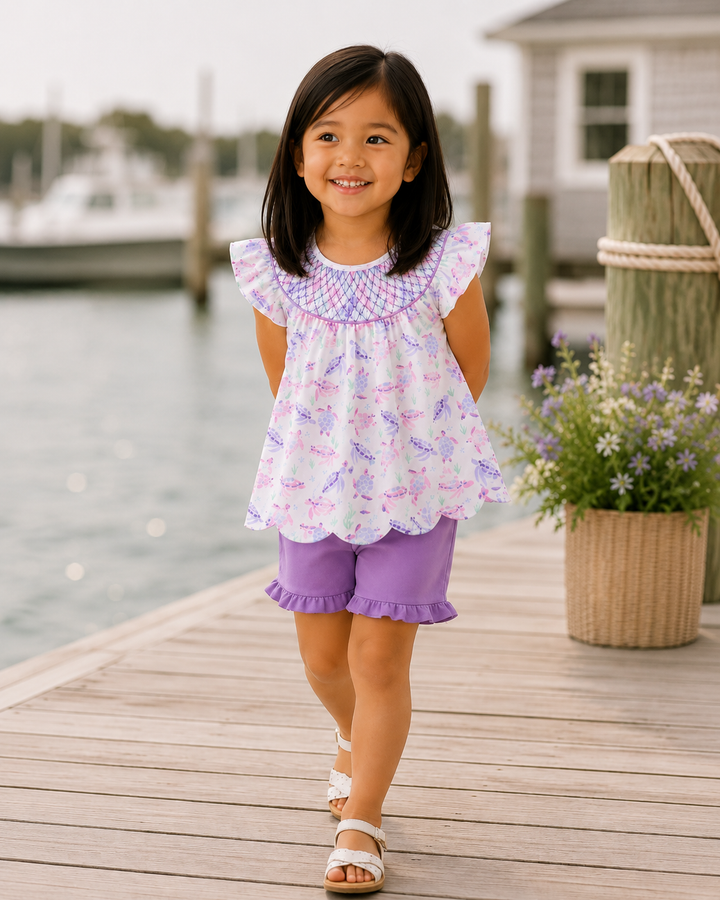 Young girl in a purple outfit standing on a dock with water and flowers in the background