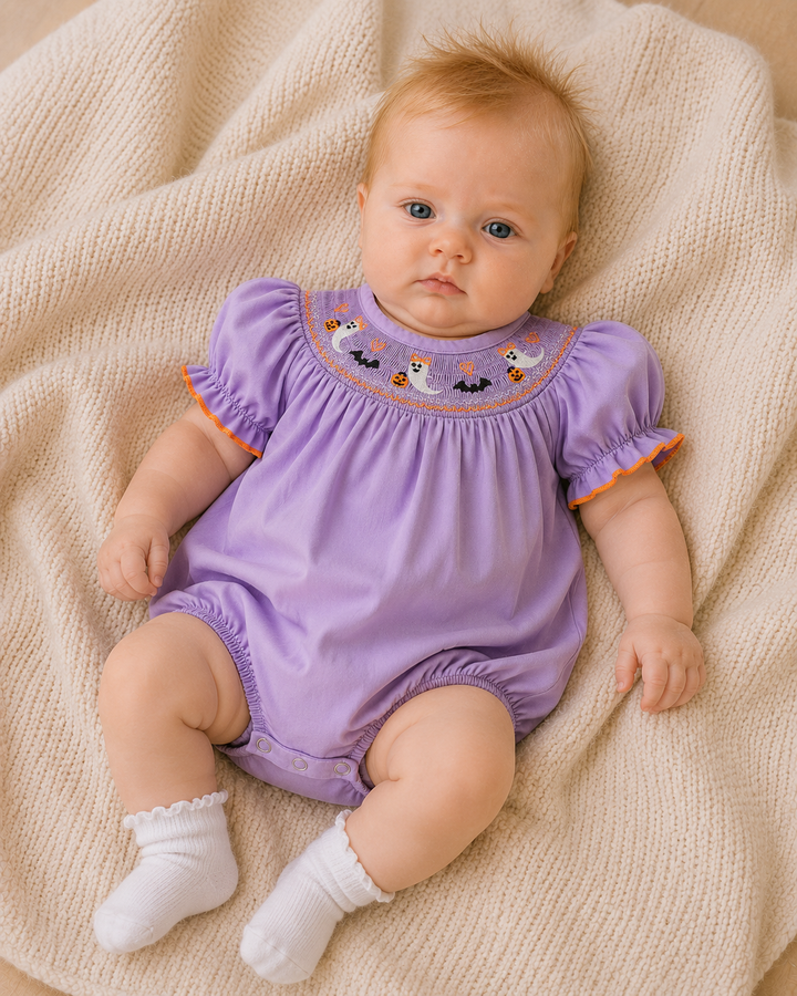 A baby lying on a cream knit blanket, wearing a lavender purple short-sleeve romper with embroidered cartoon characters around the neckline and white socks.