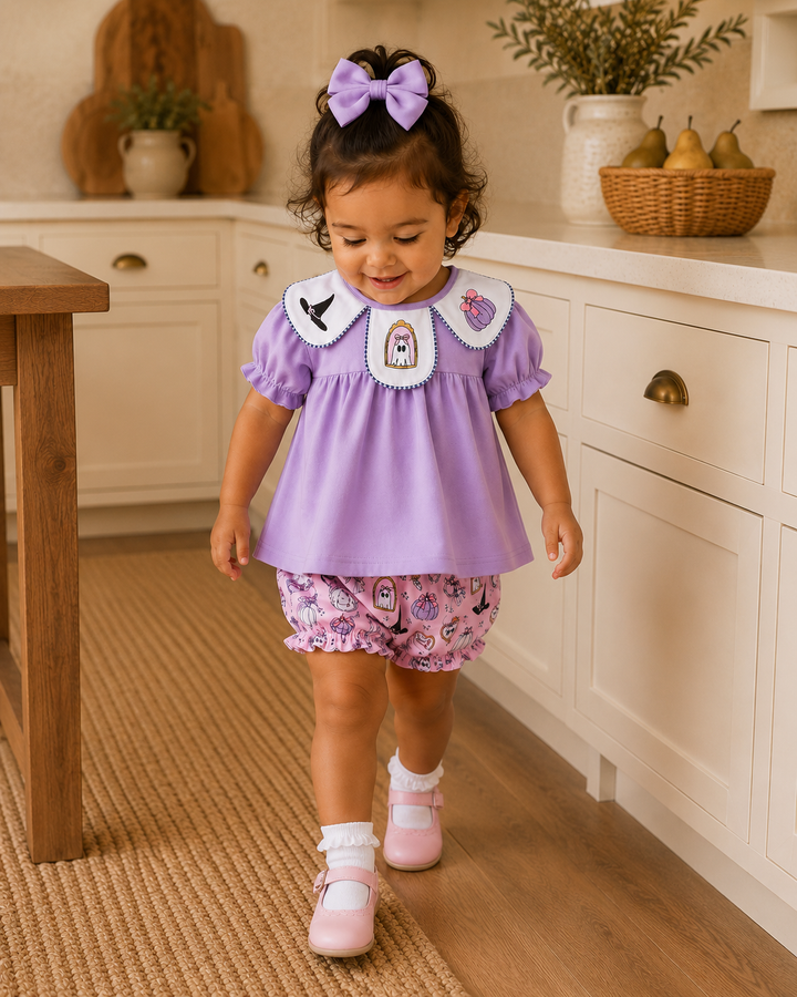 A toddler girl walking in a bright kitchen, wearing a lavender short-sleeve top with a white Peter Pan collar and embroidered details, matching floral print bloomers, white socks, and pink shoes.