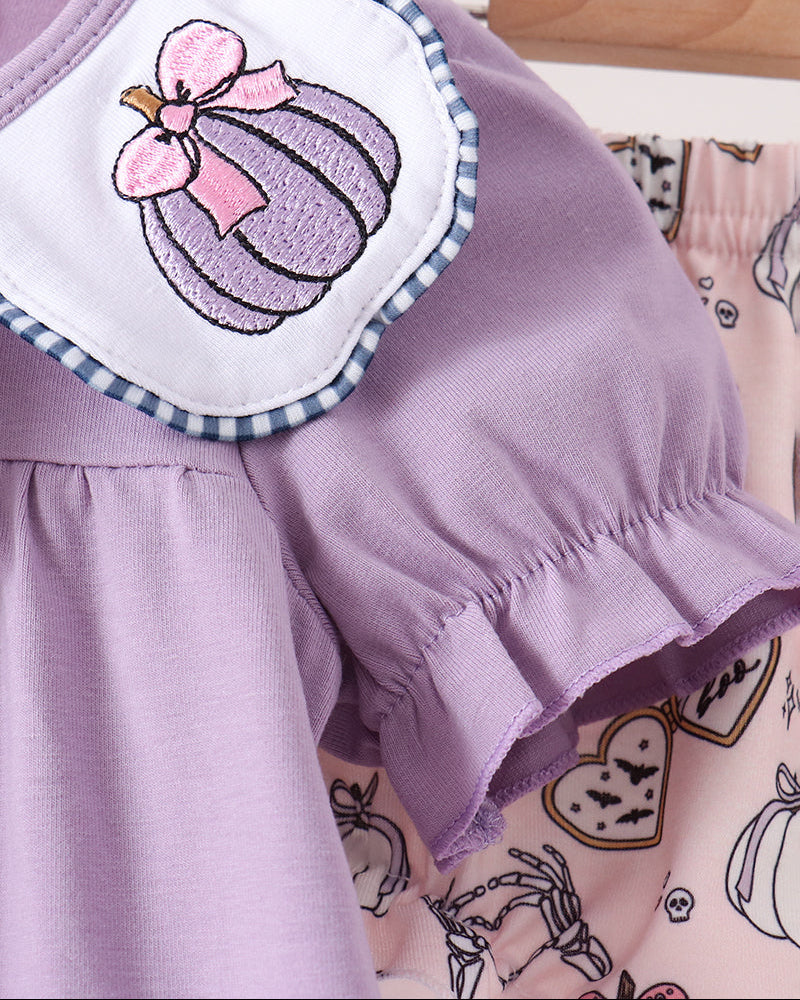 A close-up of a baby clothing set featuring a lavender short-sleeve top with a white bib collar decorated with an embroidered purple pumpkin and pink bow, hanging next to a pink patterned romper.