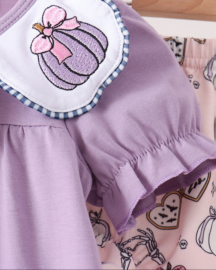 A close-up of a baby clothing set featuring a lavender short-sleeve top with a white bib collar decorated with an embroidered purple pumpkin and pink bow, hanging next to a pink patterned romper.