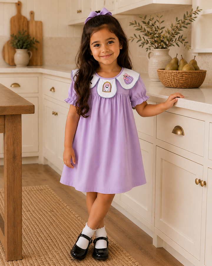 A young girl with long dark hair stands in a bright kitchen wearing a lavender dress with a white Peter Pan collar featuring small embroidered character details. She is smiling and leaning on a white countertop with wooden cabinets, a woven rug, and pears in the background.