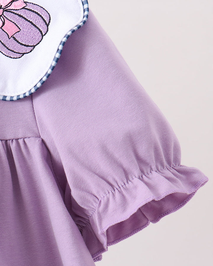Close-up of a lavender-purple dress with a ruffled sleeve, gathered waist seam, and a white collar featuring a pink pumpkin graphic and blue gingham trim.