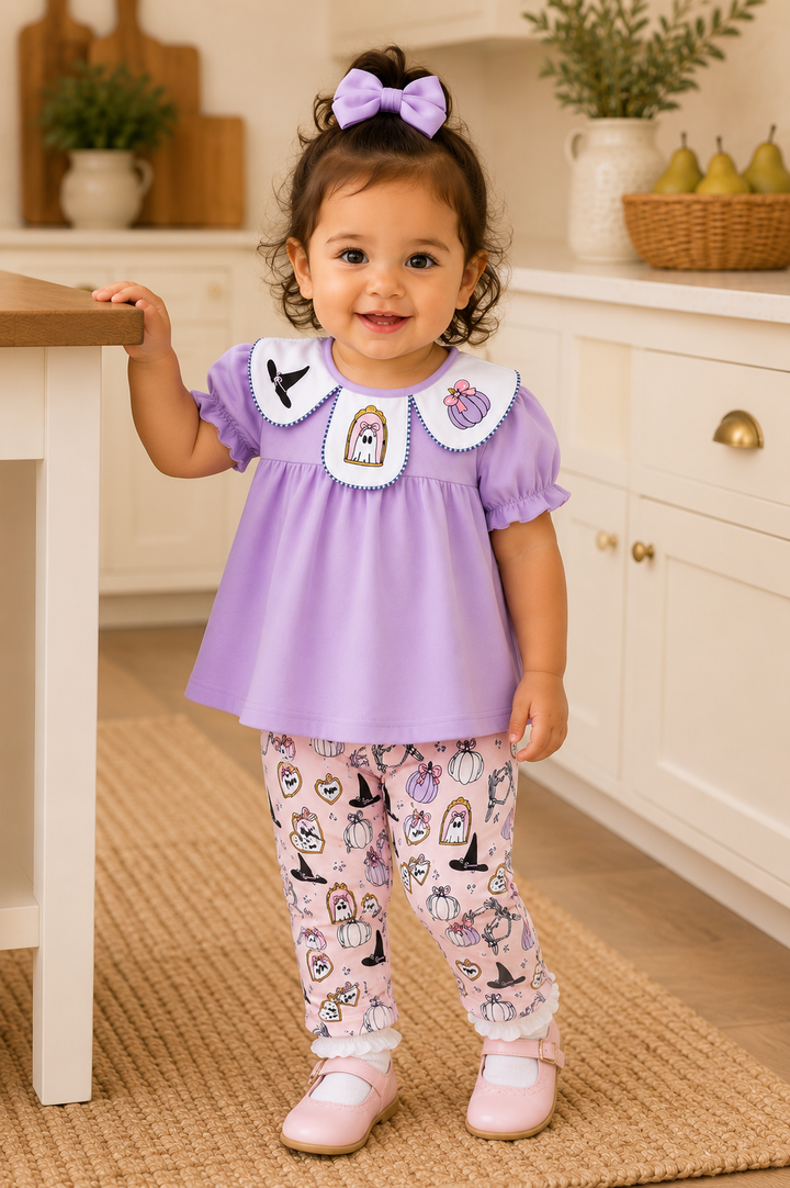 A toddler girl wearing a lavender short-sleeve dress with a white Peter Pan collar and a purple bow in her hair, paired with pink patterned leggings and light pink shoes, standing in a bright kitchen setting.