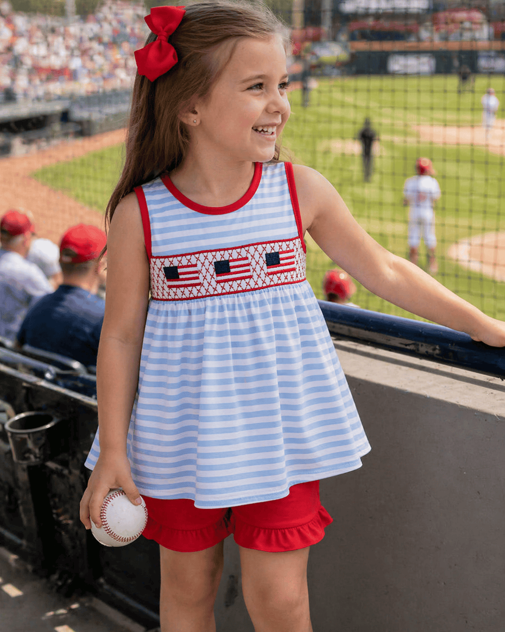 Young girl in a striped dress with American flag design at a baseball game.
