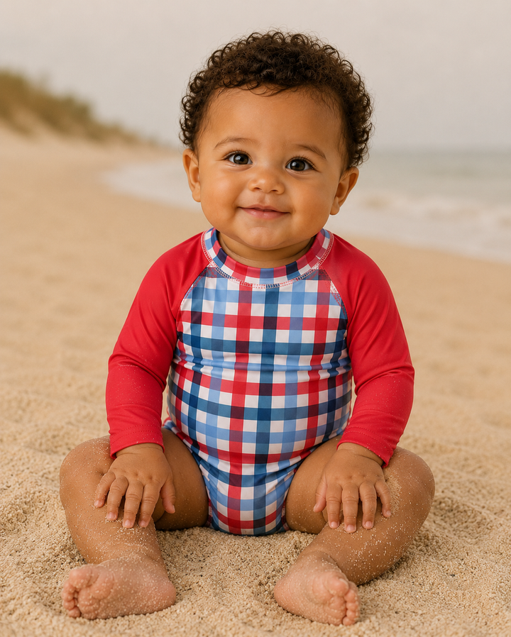 Baby wearing a red and blue checkered swimsuit on a sandy beach.