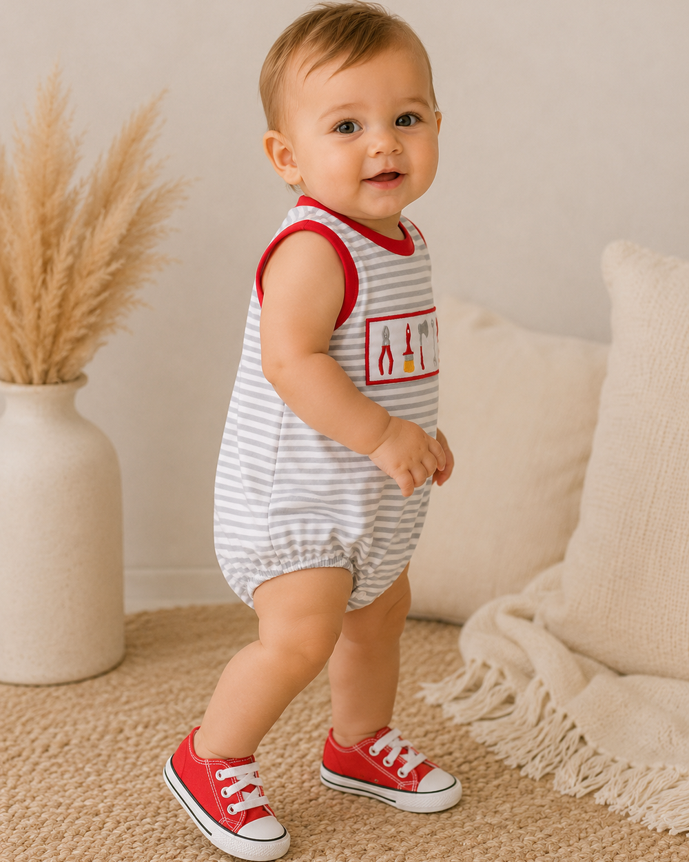 A baby toddler wearing a sleeveless white and gray striped romper with red trim and a red chest graphic, paired with red sneakers, standing in a cozy room setting.