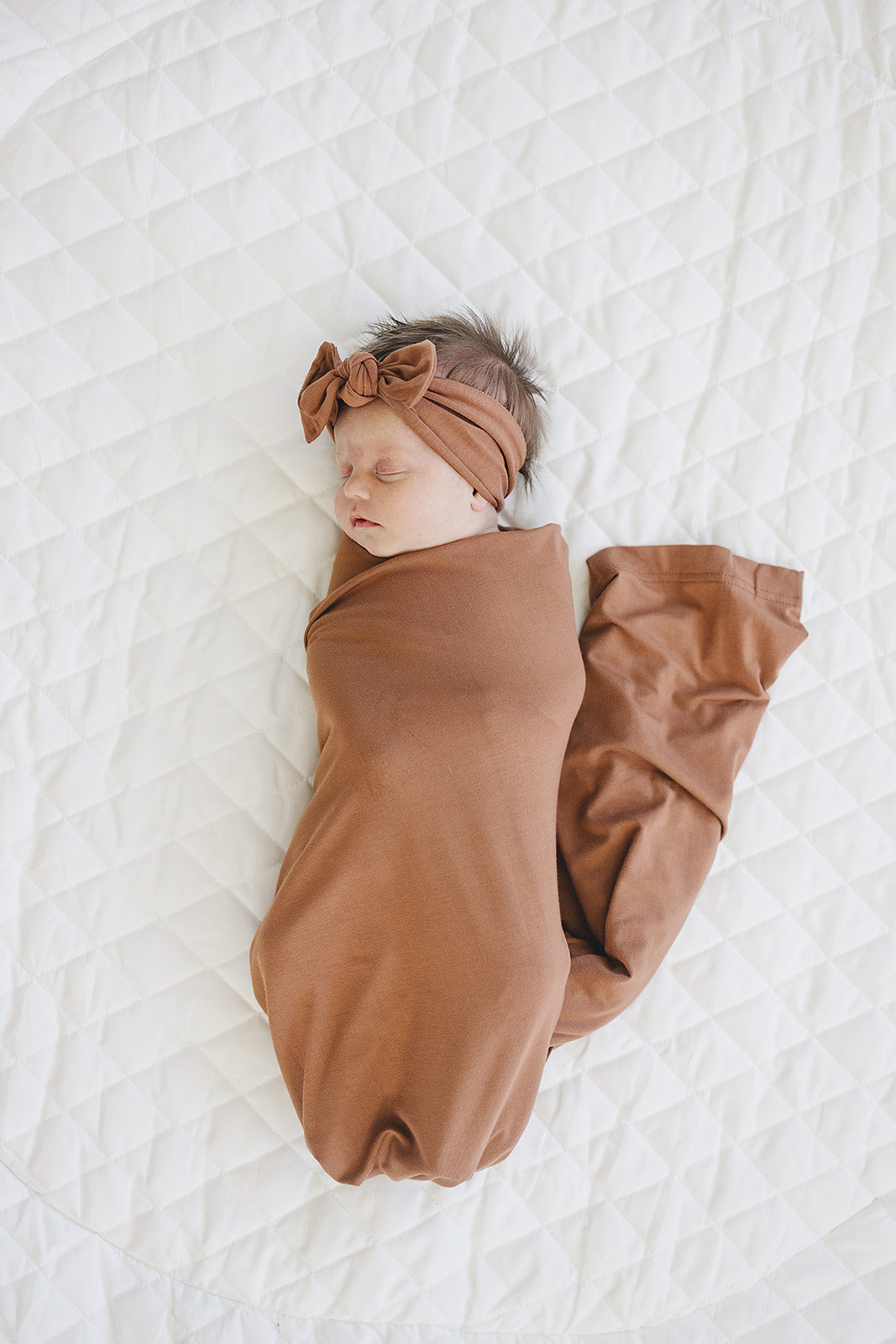 Rust knotted head wrap with bow on a baby wrapped in a brown swaddle