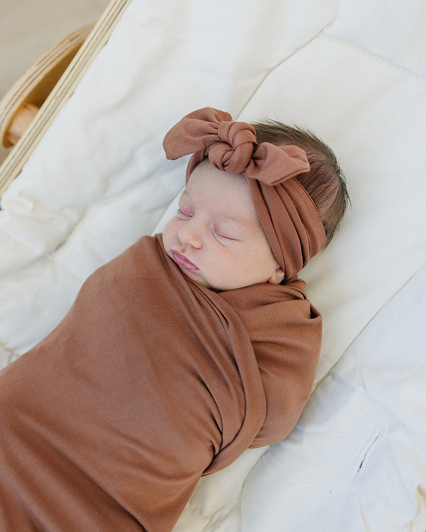 Rust knotted bamboo head wrap with bow on a baby lying on white bedding