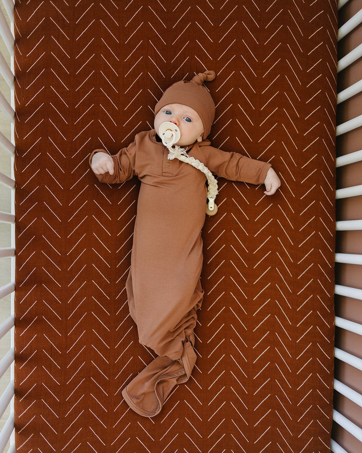 Baby in a knot gown with a hood lying on a brown blanket with a white zigzag pattern.