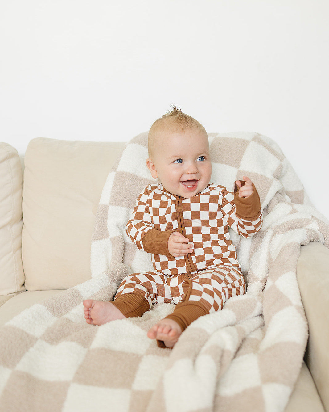 Baby in a checkered outfit sitting on a couch with a blanket